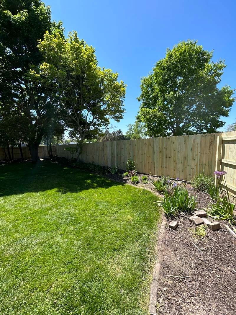 Backyard with a wooden fence, green grass, and a flower bed under a clear blue sky.