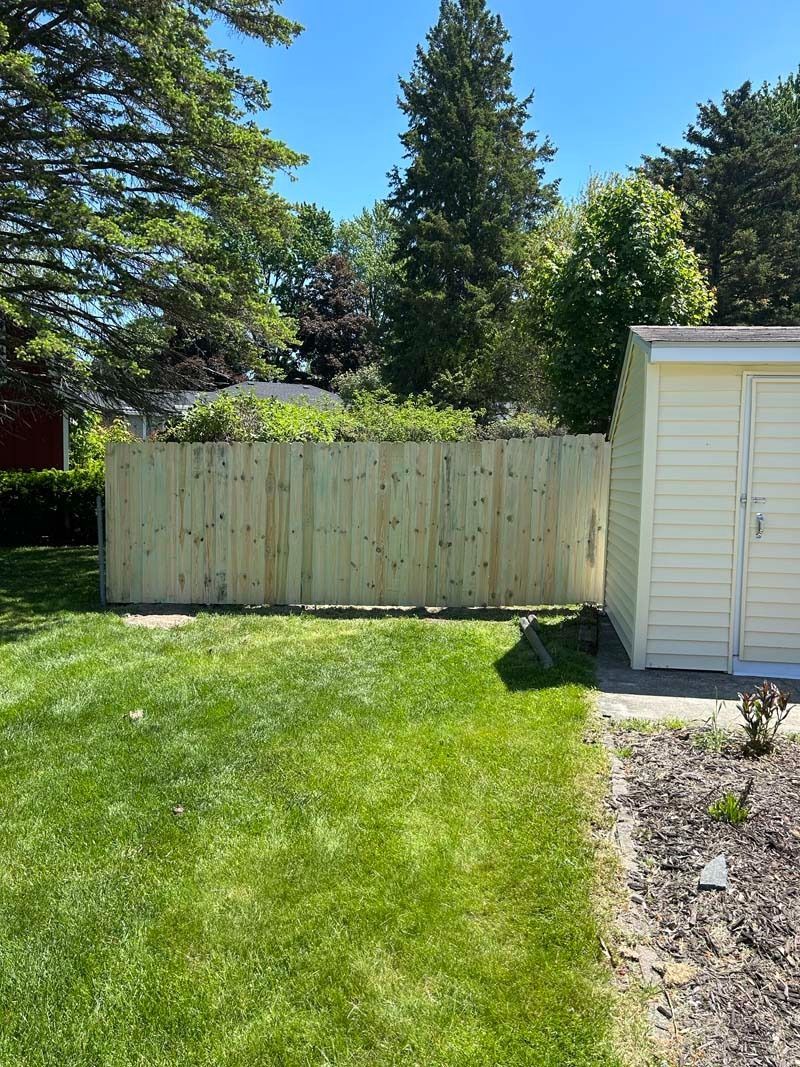 Wooden fence next to a light yellow shed on a grassy lawn under a sunny sky.