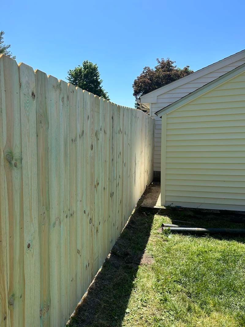 Wooden fence alongside a yellow house and green grass under a clear blue sky.
