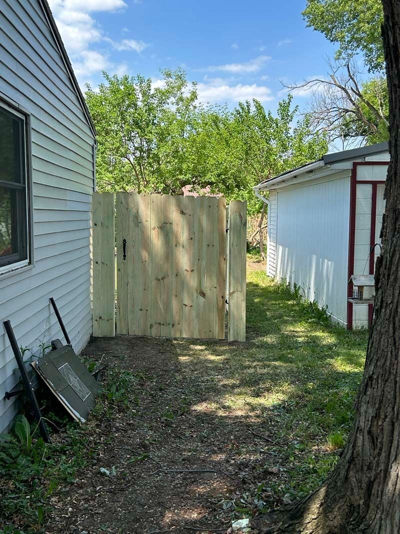 Wooden fence blocks a narrow backyard path between a house and shed on a sunny day.