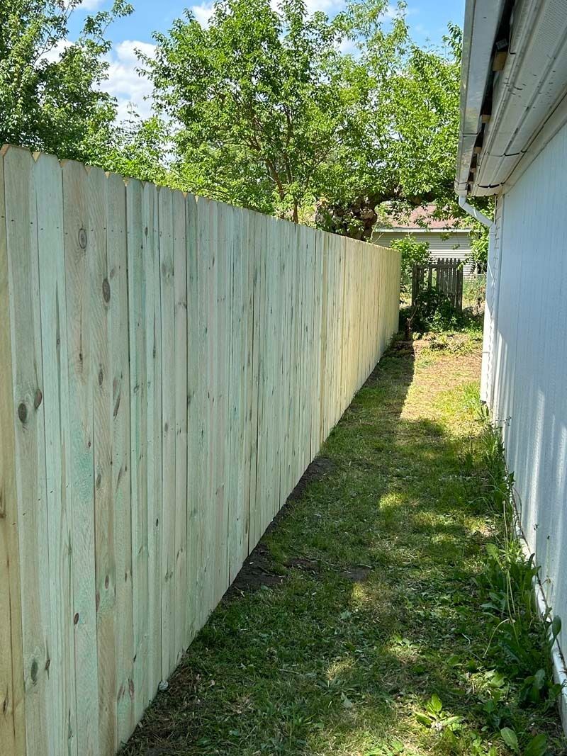 Wooden fence alongside a white house, with a narrow grassy space in between.