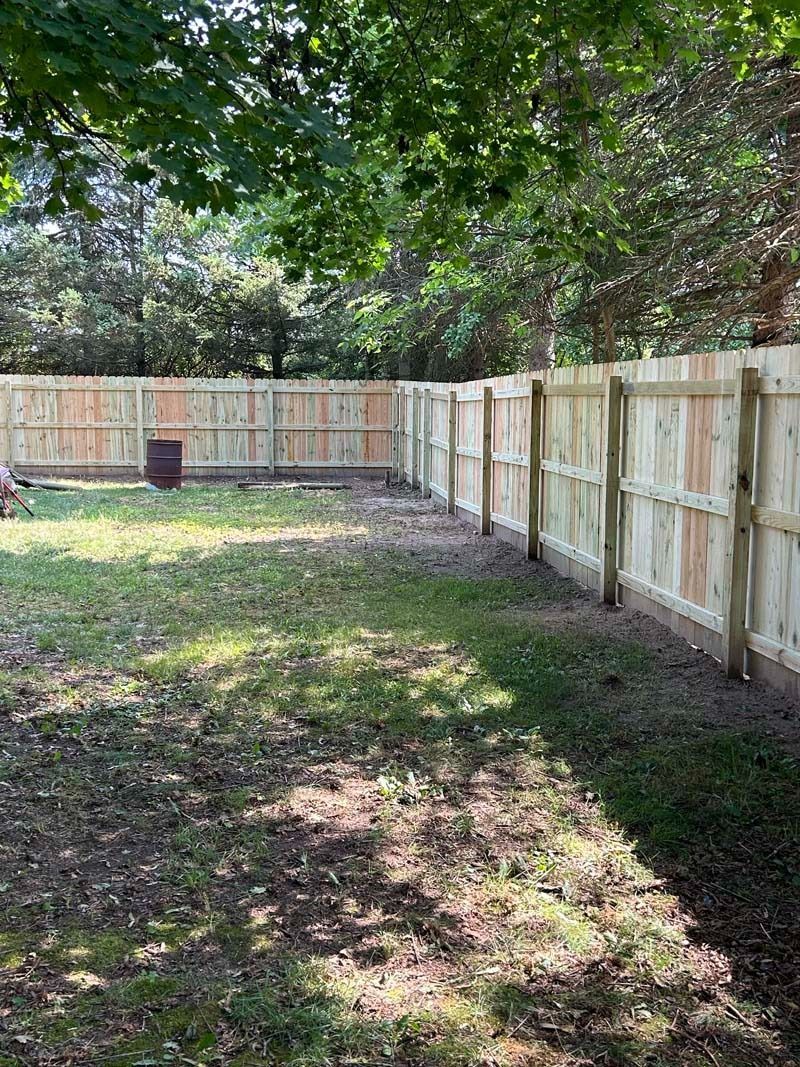 Backyard with a wooden fence, green grass, and trees in the background on a sunny day.