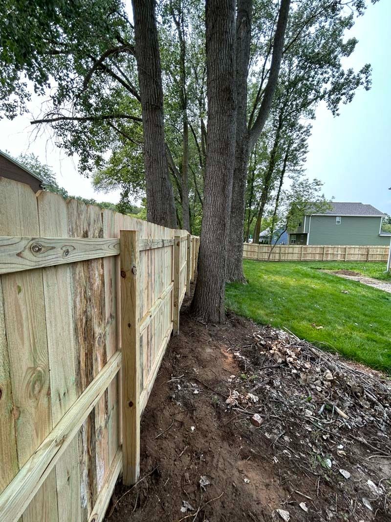 Wooden fence running next to trees on a slight slope with a green yard and houses in background.