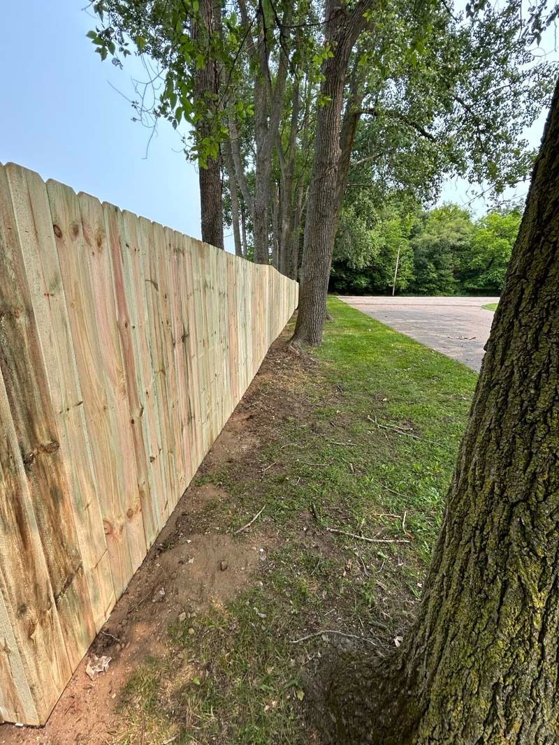 Wooden fence bordering a grassy yard with trees and a paved road in the background.