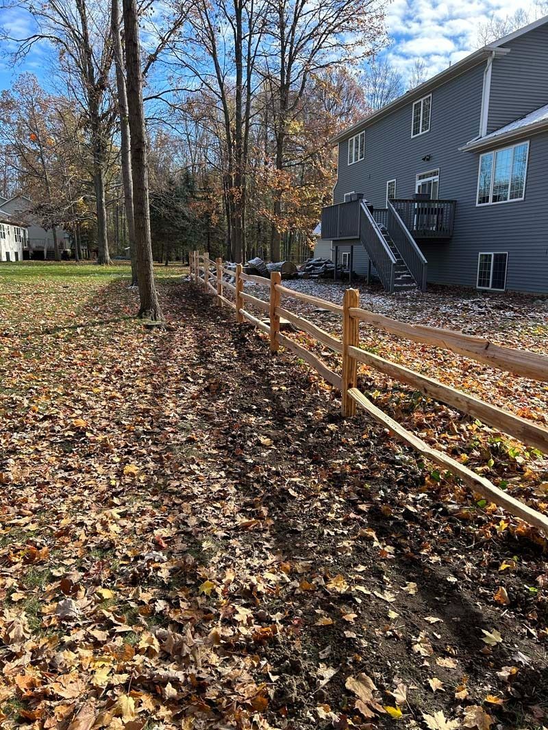 A split-rail fence borders a backyard with fallen leaves and a house with a deck.