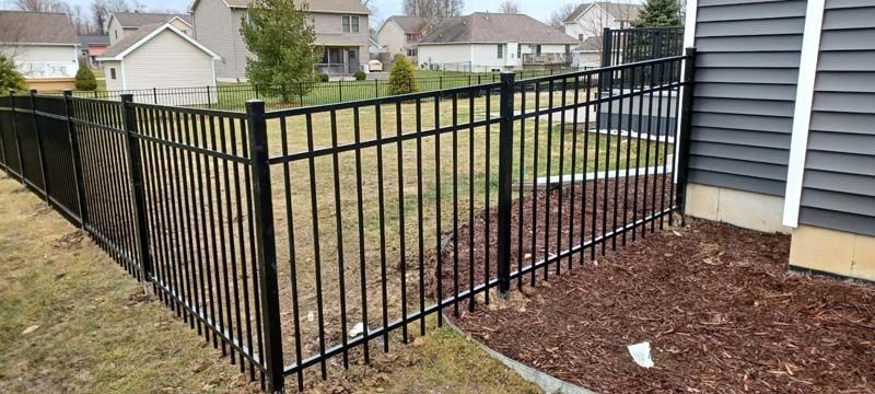 Black metal fence in front of a house with gray siding, and a backyard with mulch.