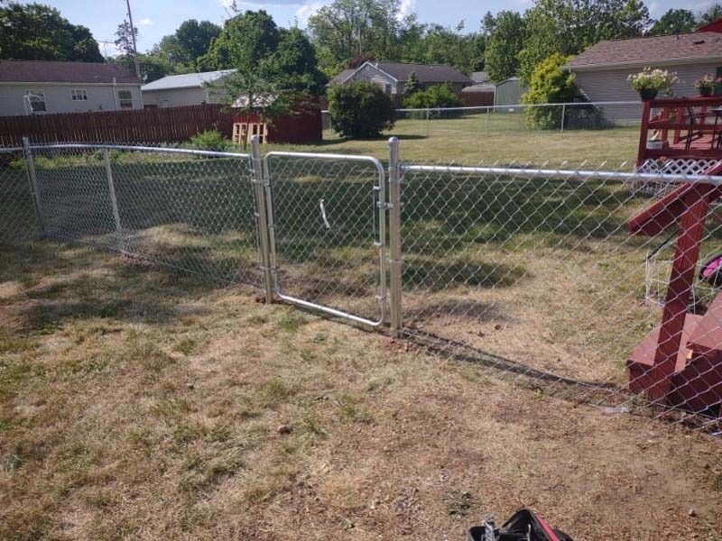 Chain-link fence with gate in a yard with brown grass, red deck, and houses in the background.