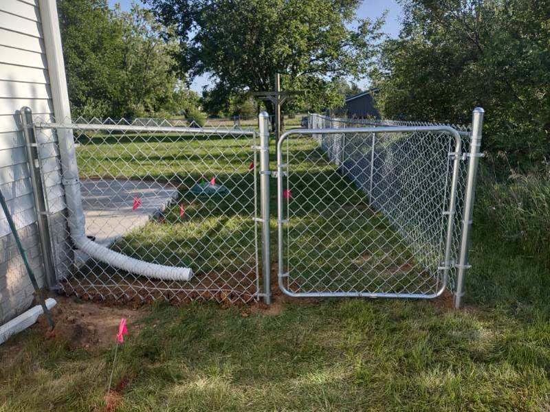 Chain-link fence with a gate by a house. Green grass, pink flags, and trees in the background.