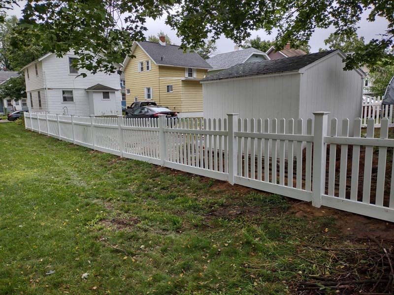 White picket fence in a grassy yard next to a white shed and several houses.