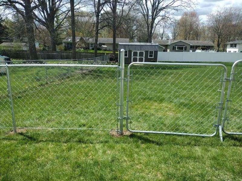 A chain-link fence encloses a grassy yard, with a gate open, houses and trees in the background.