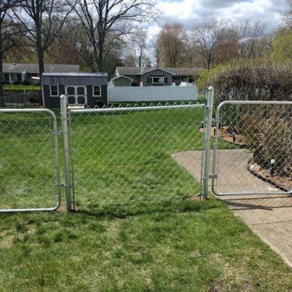 Chain-link fence with two open gates. A shed and houses are visible in the background. Green grass surrounds the fence.