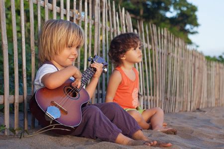 Two little boys sit on the beach playing ukulele