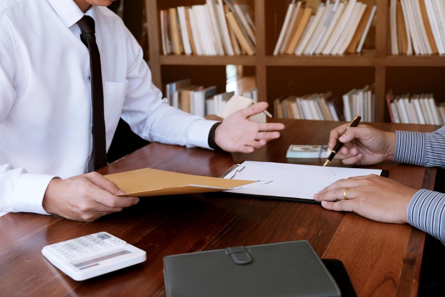 Two people at a wooden desk with papers, a calculator, and a file. One gestures while the other signs.