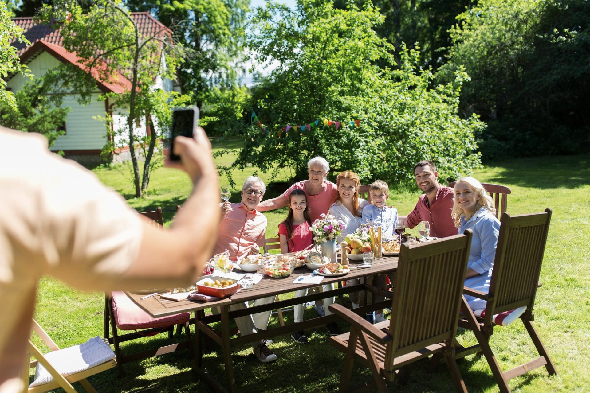 Person takes photo of family gathered around a table outdoors, set for a meal.