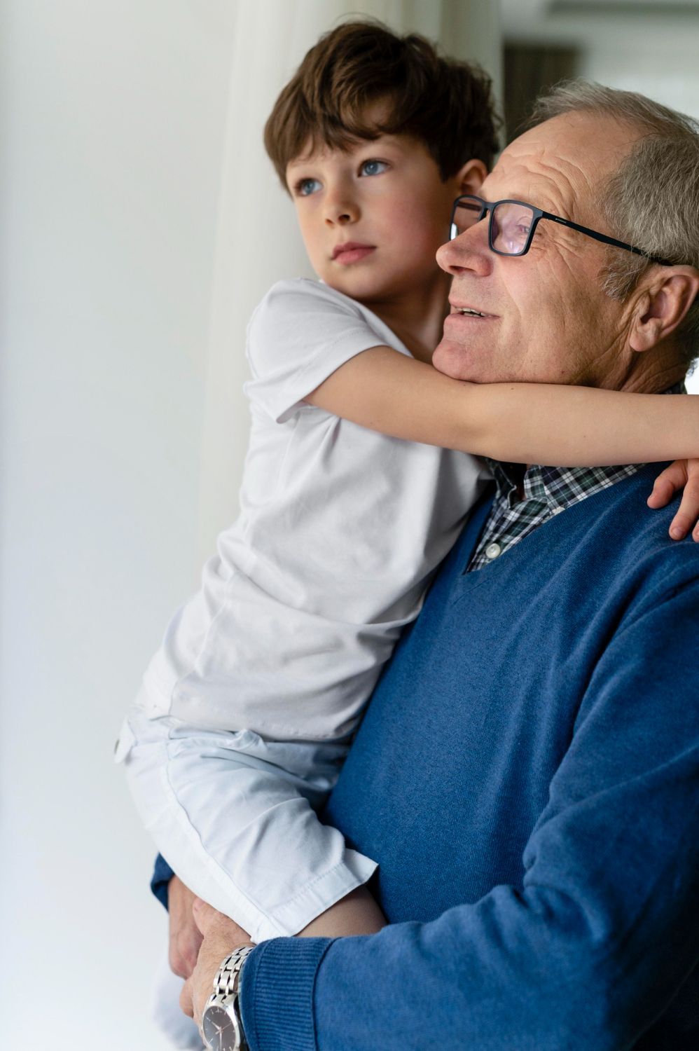 Boy hugging older man, both looking toward a window. Man wearing glasses and blue sweater.
