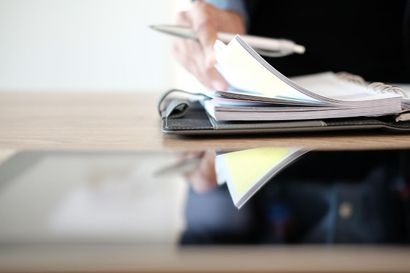 Person flipping through pages in a notebook, pen in hand, near a tablet on a desk.