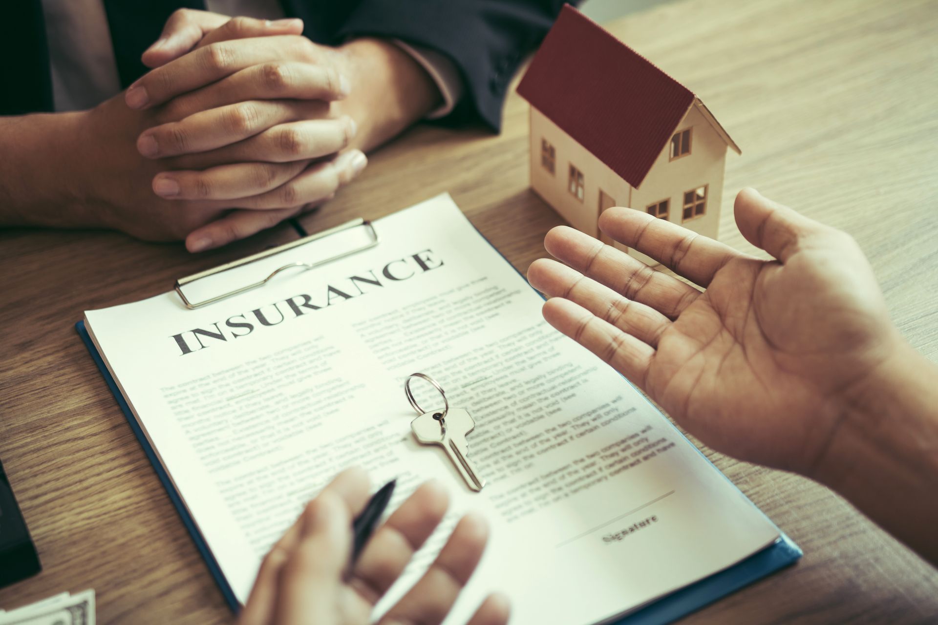 Hands gesturing over an insurance document with keys and a miniature house on a desk.