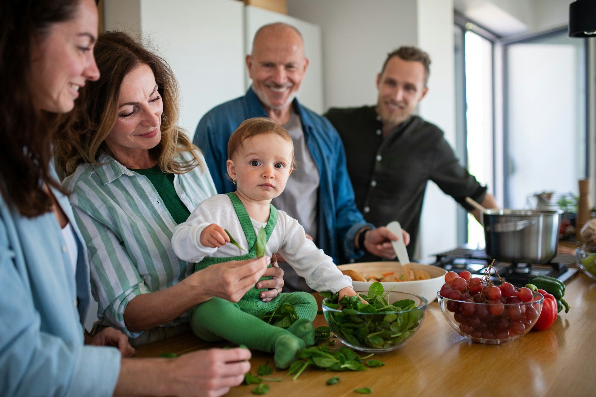 Family cooking together in a kitchen, baby held in arms, food on the counter, people smiling.