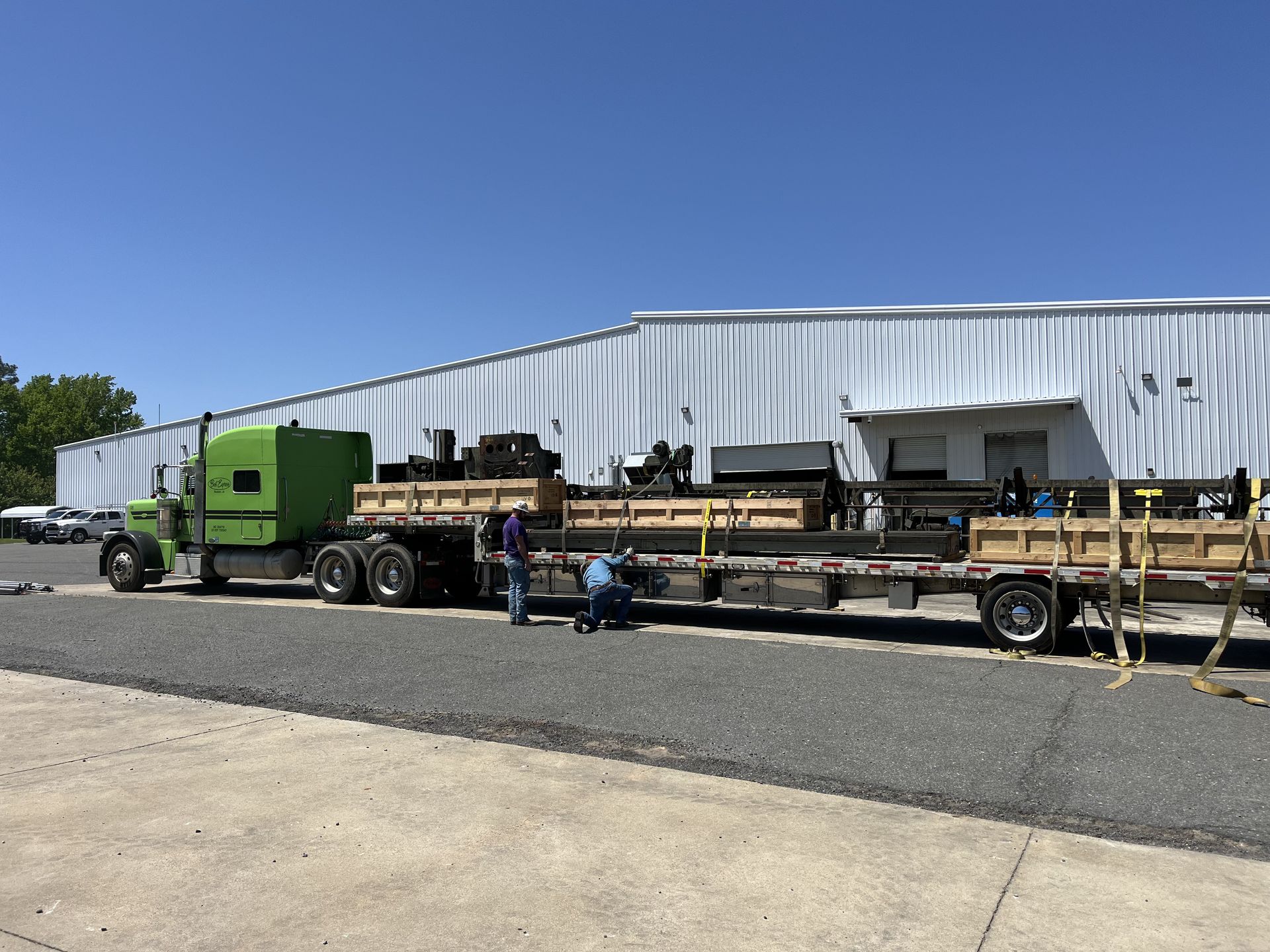 Green semi-truck loaded with machinery, two people securing cargo outdoors in front of a white building.