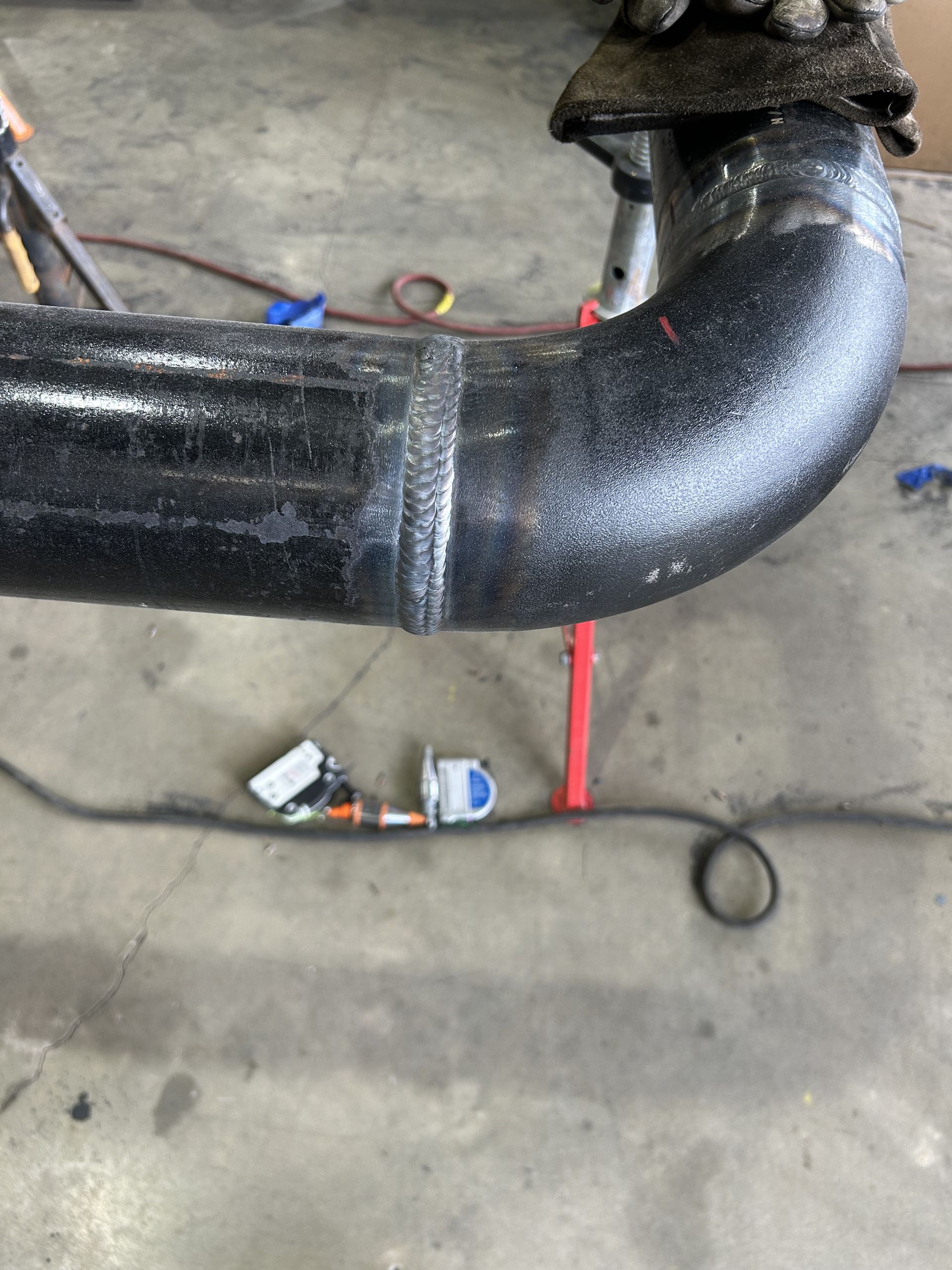 Black pipe elbow with welded joint, resting on a concrete floor with tools in the background.