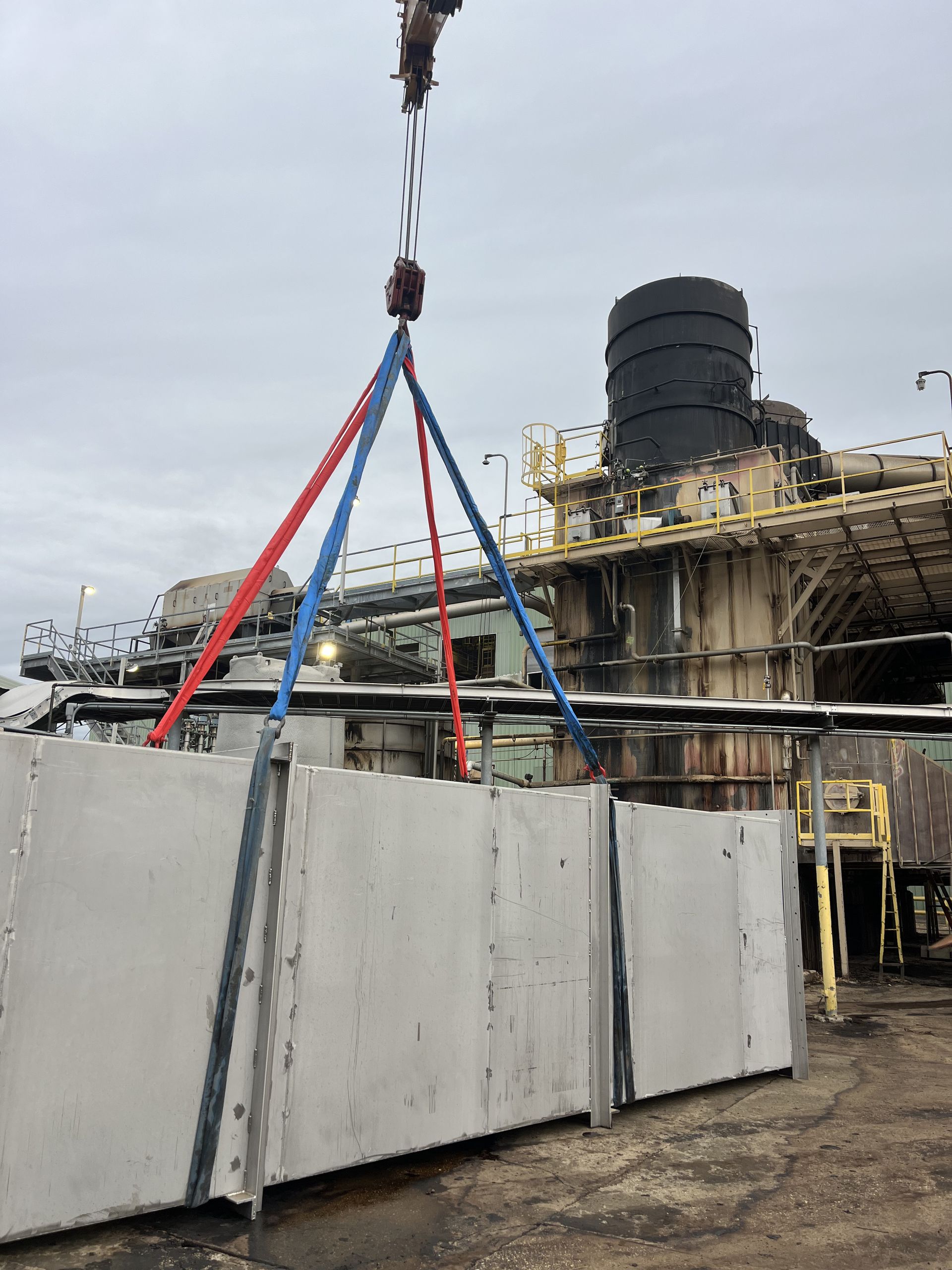 Crane lifting concrete panels at industrial site. Red and blue straps secure the panels.