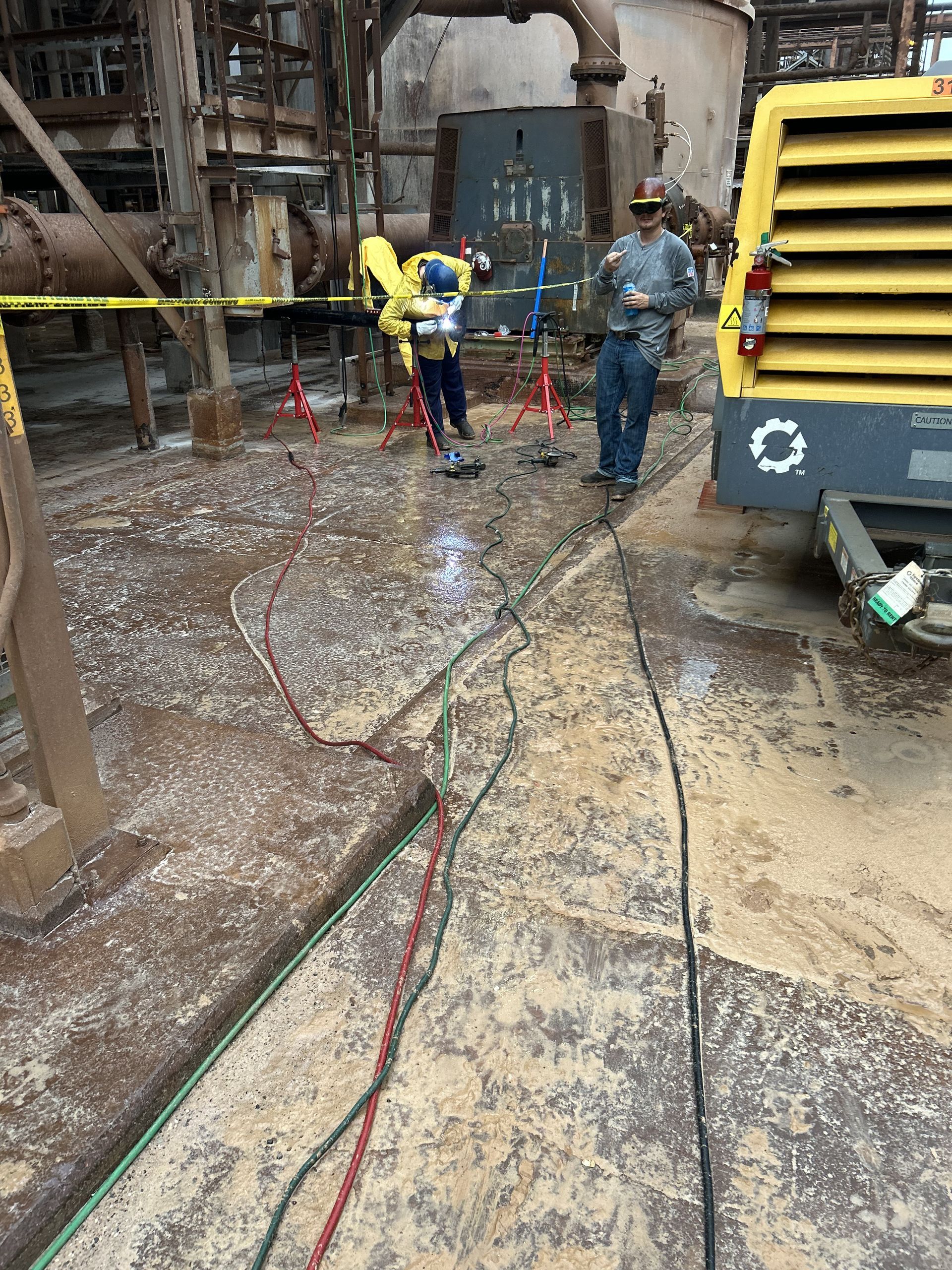 Two workers in an industrial setting. One operates a saw, the other watches. Equipment and lines are on the wet concrete floor.