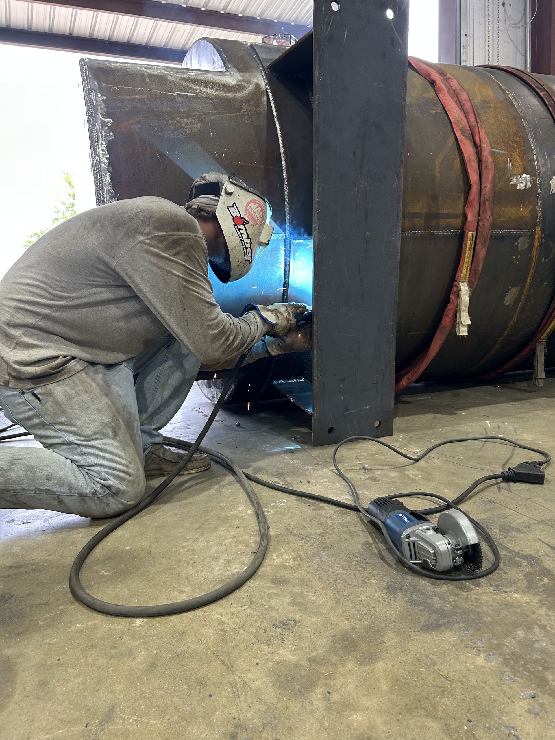 Welder kneeling, welding metal structure. Protective mask and gloves visible. Sparks fly.