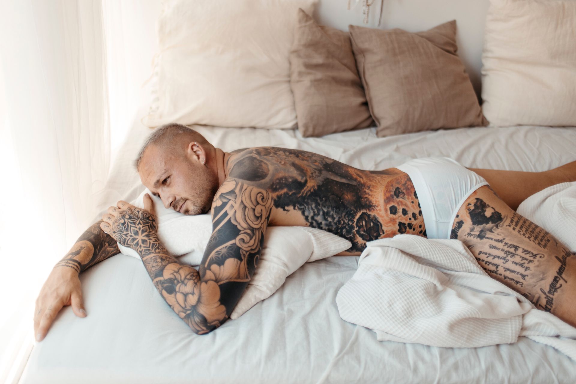 Uomo con tatuaggi sdraiato su un letto, con indosso slip bianchi, appoggiato su un cuscino e con lo sguardo rivolto di lato.