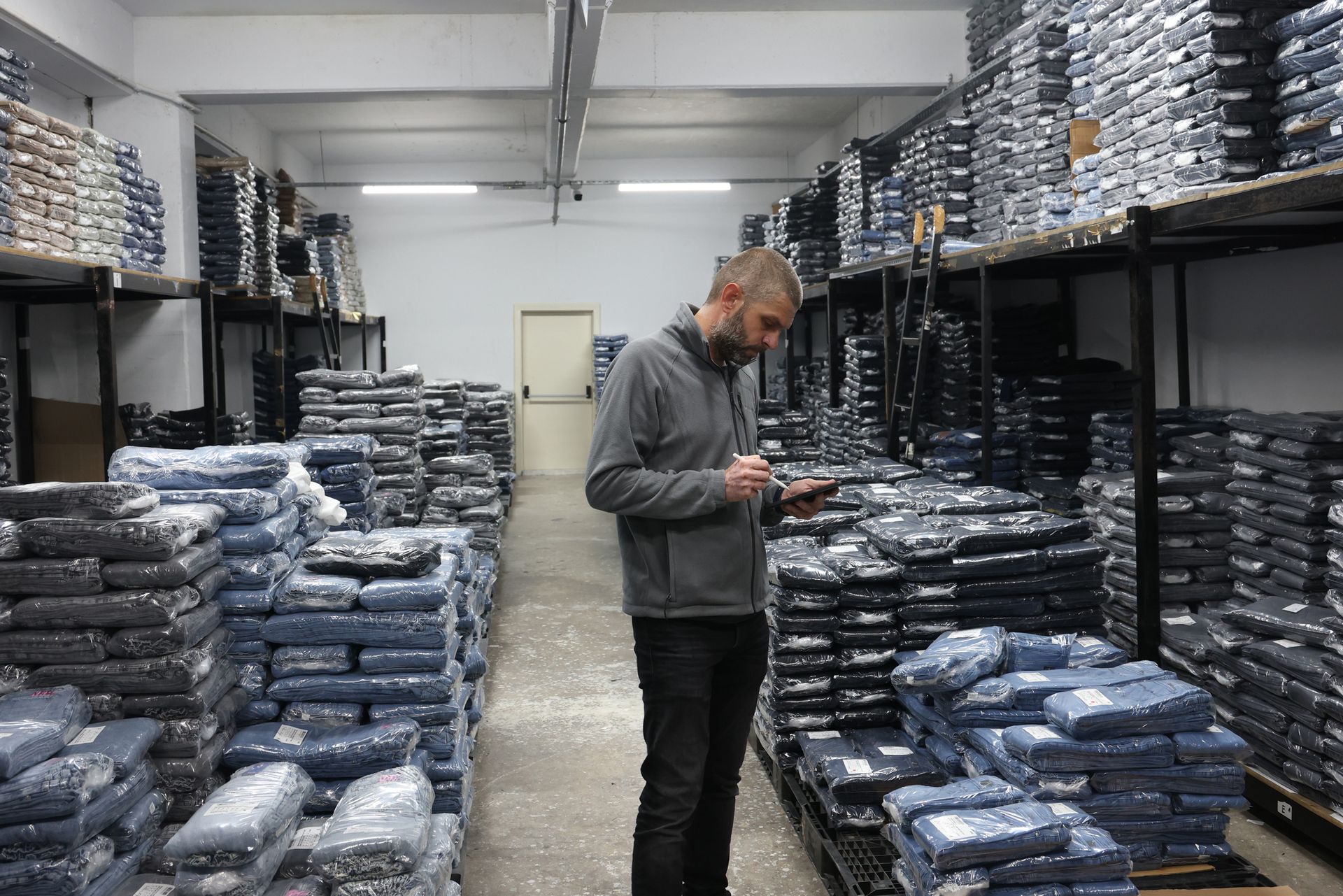 Un uomo vestito di grigio esamina un tablet in un magazzino pieno di pile di articoli confezionati di colore scuro.