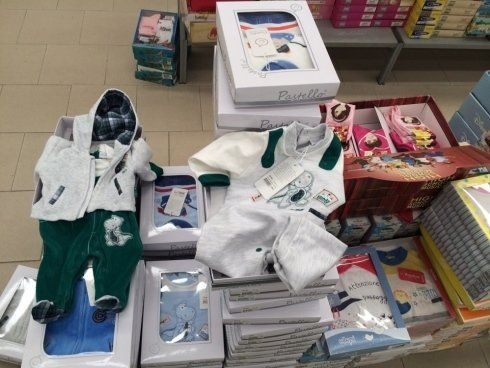Vestiti per neonati esposti sugli scaffali di un negozio, tra cui completini, in vari colori e confezioni.