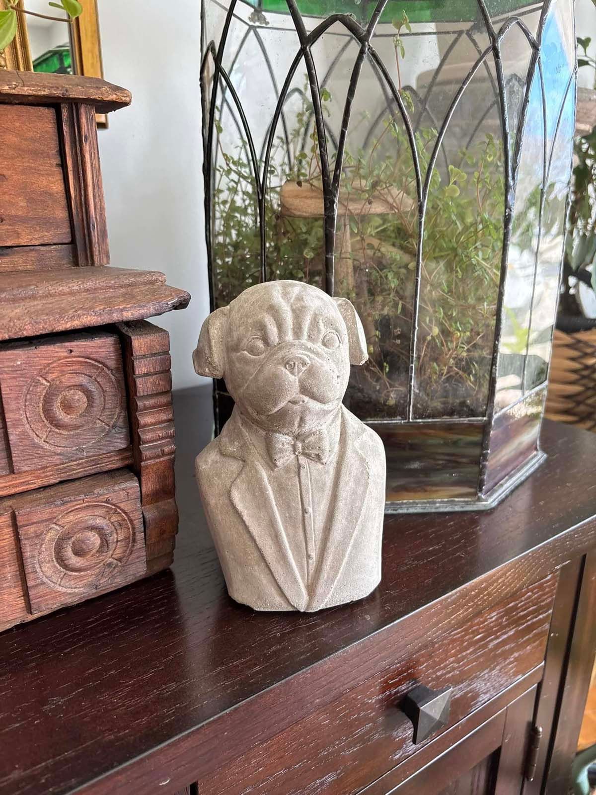 A statue of a pug wearing a suit and bow tie is sitting on a wooden dresser.