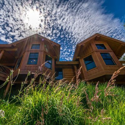 Two-story wooden cabin with multiple windows on a grassy hillside under a cloudy blue sky.