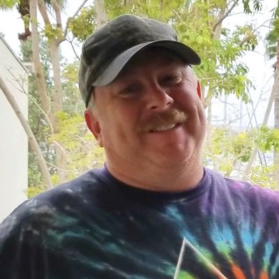 Man with mustache wearing tie-dye shirt and camo cap, smiling outdoors.