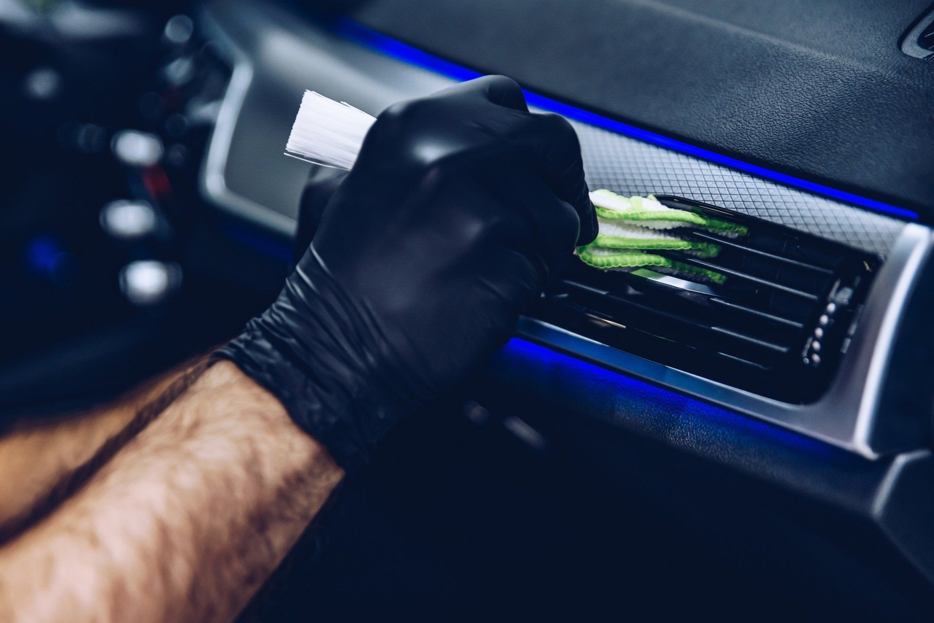 A person wearing black gloves is cleaning the air vents of a car with a brush.