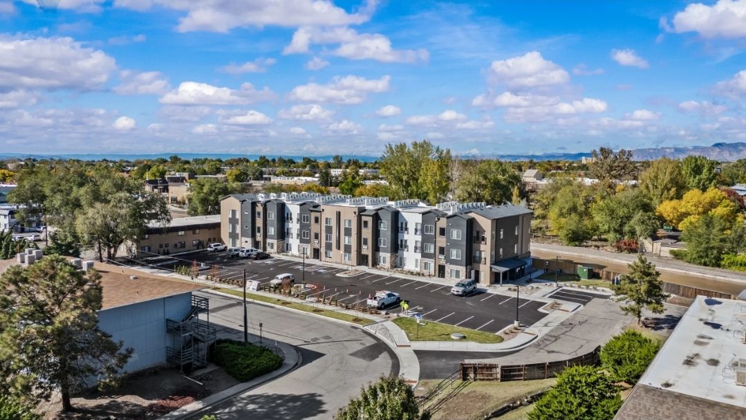 Aerial view of multi-story apartment building with parking, trees, and cloudy sky.