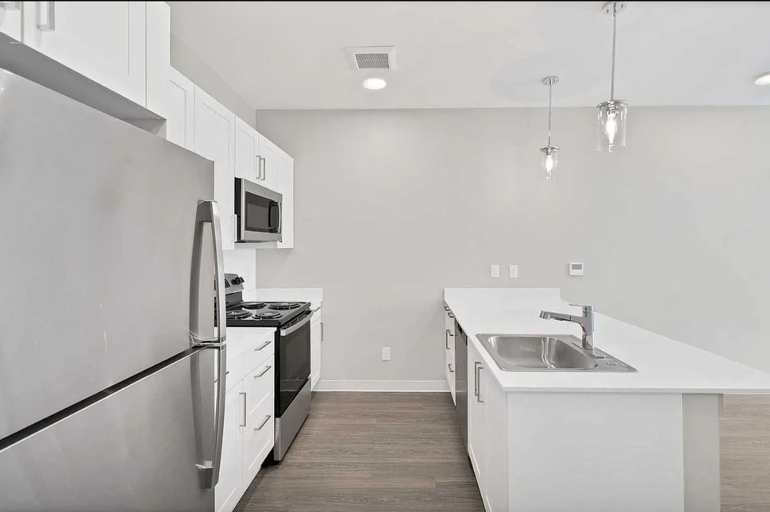 Modern white kitchen with stainless steel appliances, white island with sink, and hanging lights.