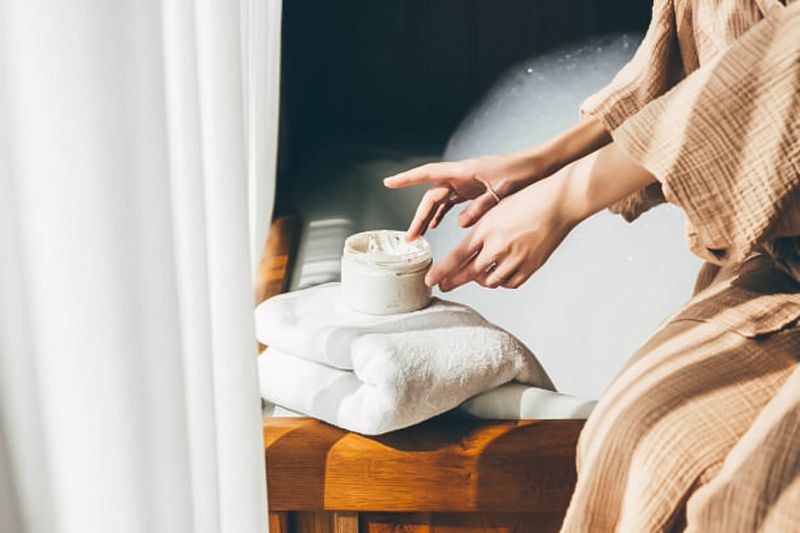 A woman is sitting on a bench in a bathtub applying cream to her hands.