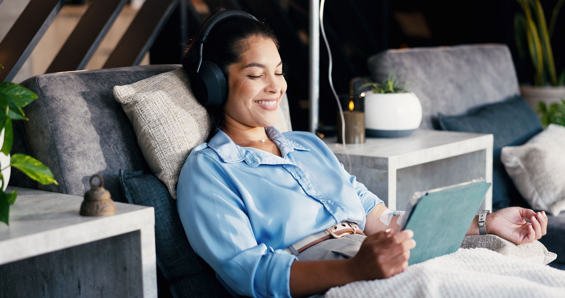 Woman relaxing on a sofa with headphones and a tablet, smiling in a cozy living room.