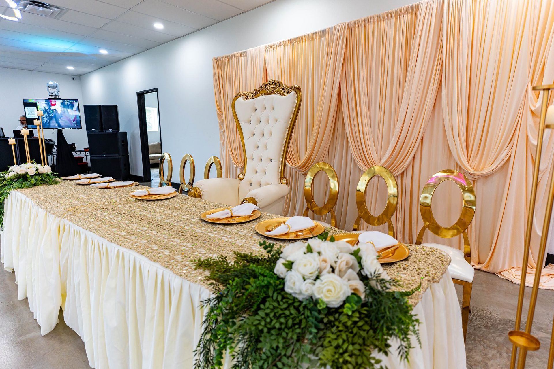 Gold-and-white decorated banquet table with ornate chair, floral centerpiece, and draped backdrop