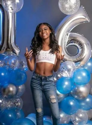 Person posing in front of blue, silver, and white balloons with a large “16” birthday display