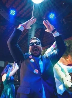 Person in a dark suit and sunglasses raises both hands on a colorful stage under bright lights