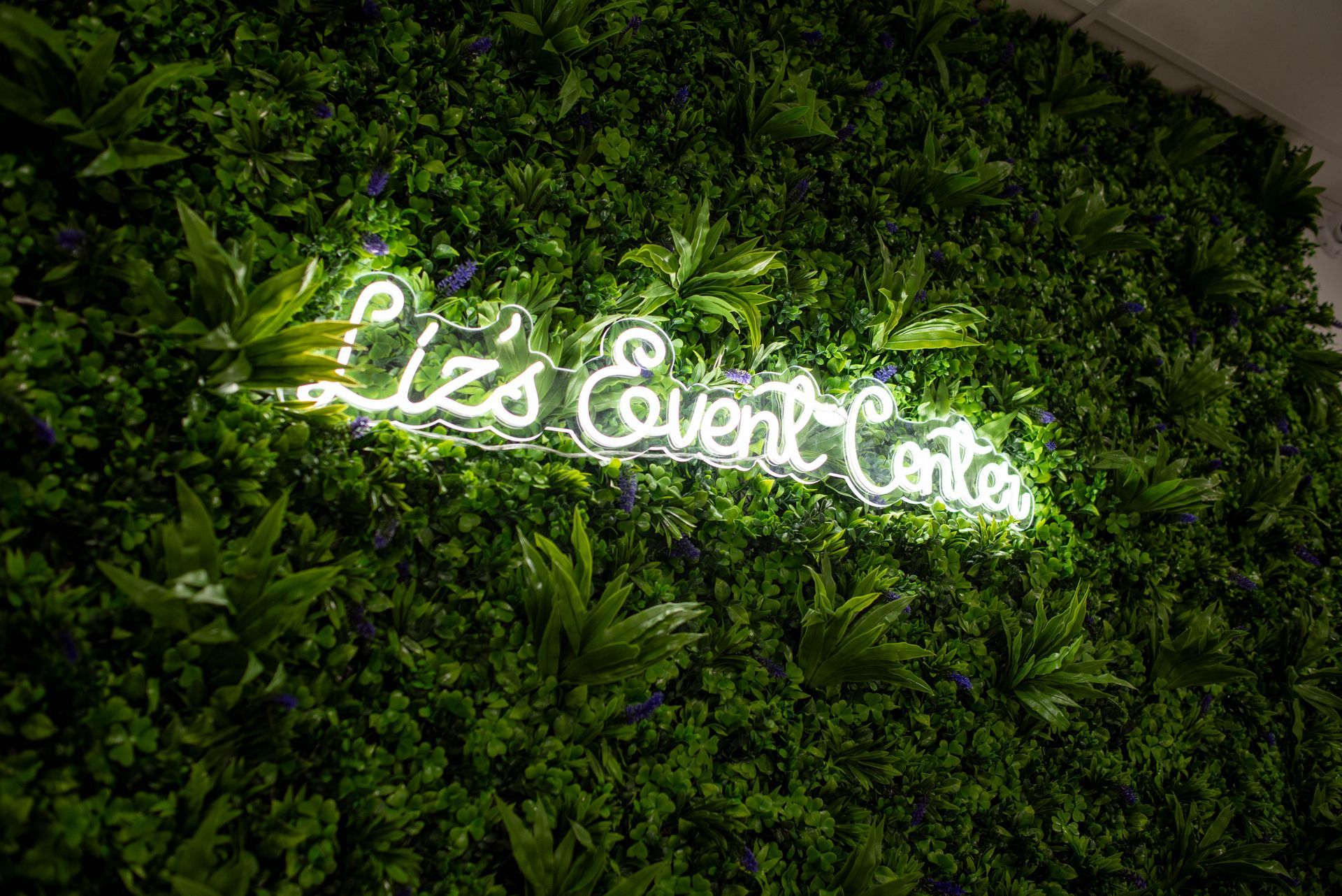 Glowing white “Liz EventCafe” sign on a lush green foliage wall