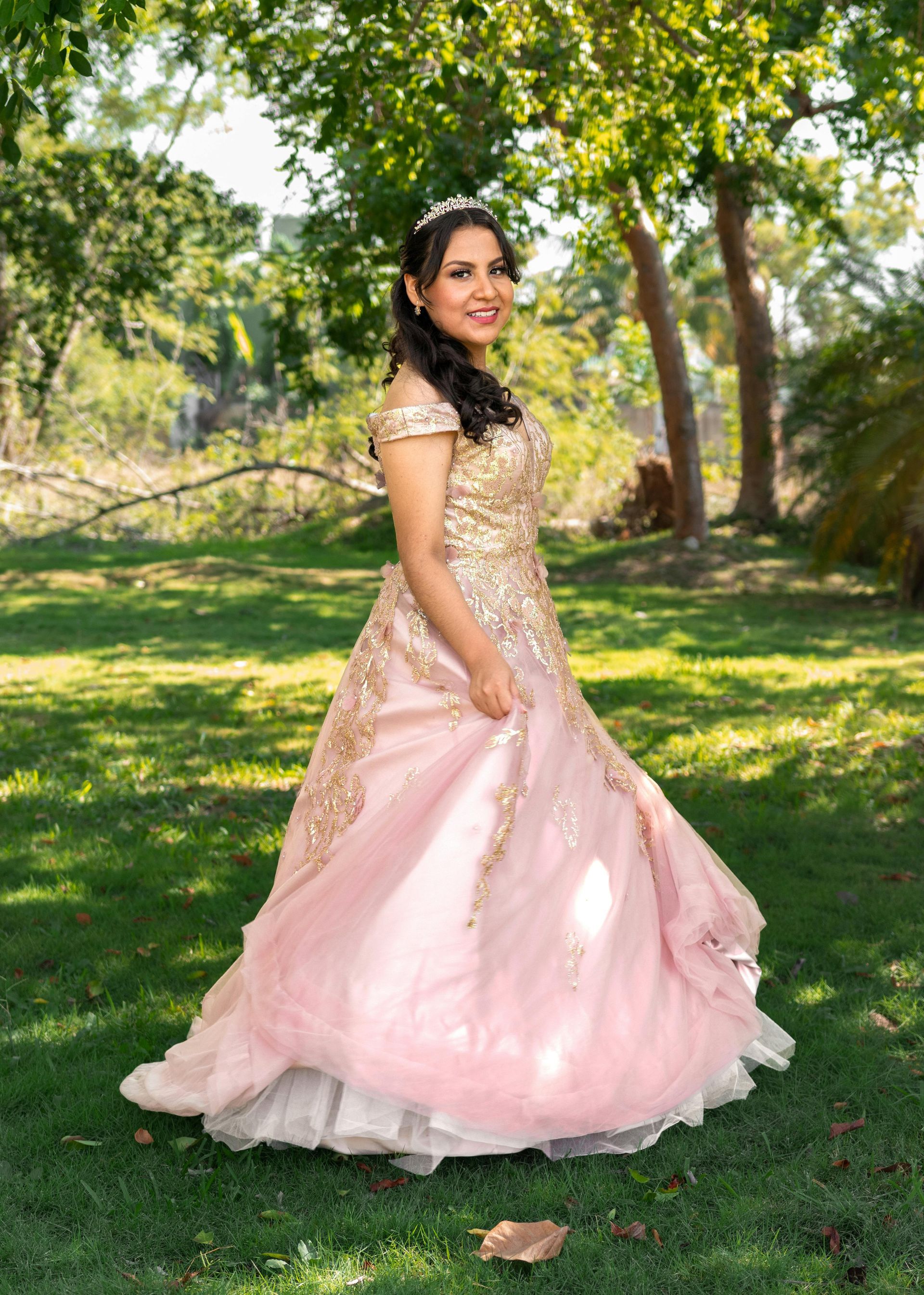 Woman in a pink gown standing on grass under trees in a sunlit park
