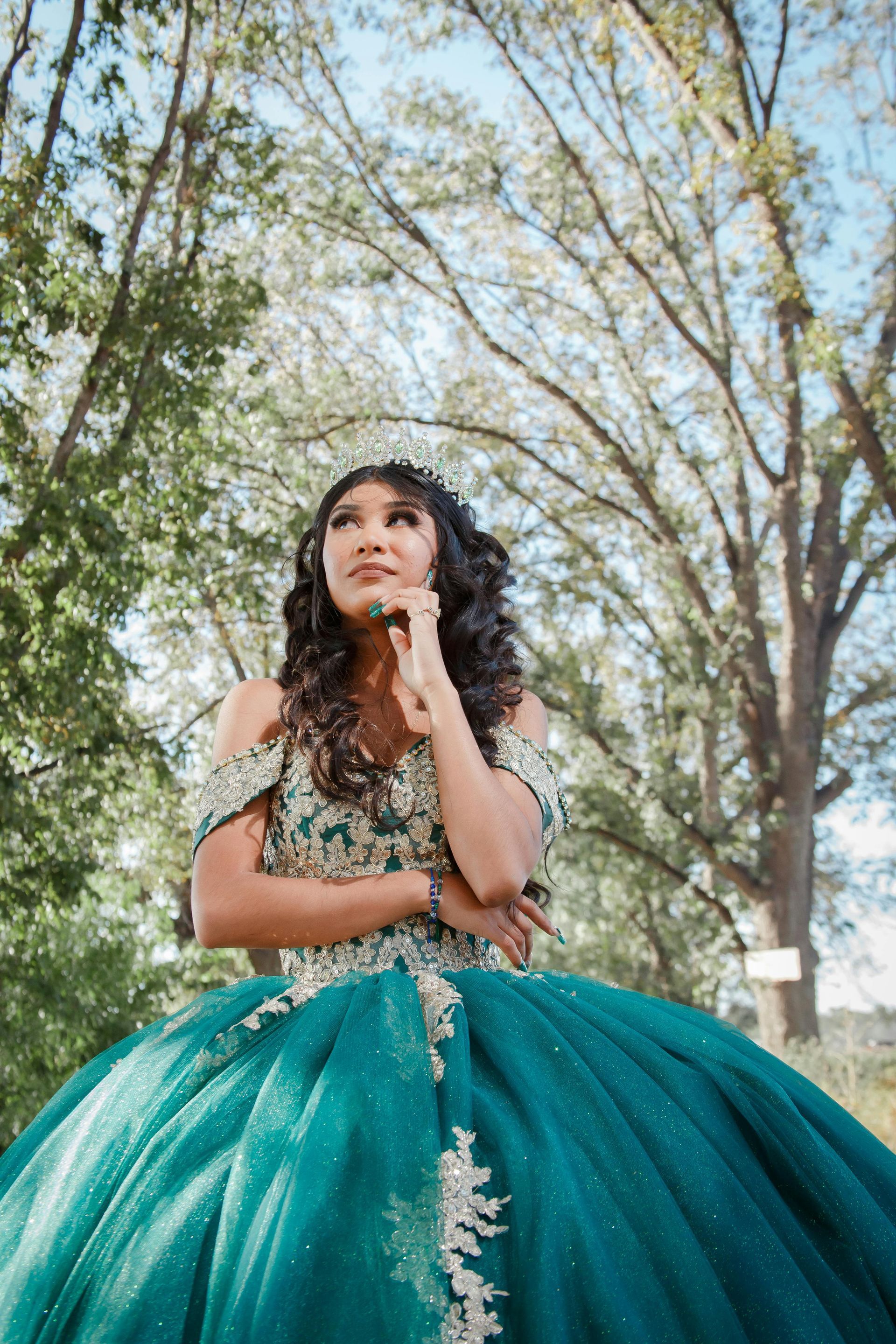Woman in a teal gown posing outdoors under tall trees, hand near chin, with a thoughtful expression