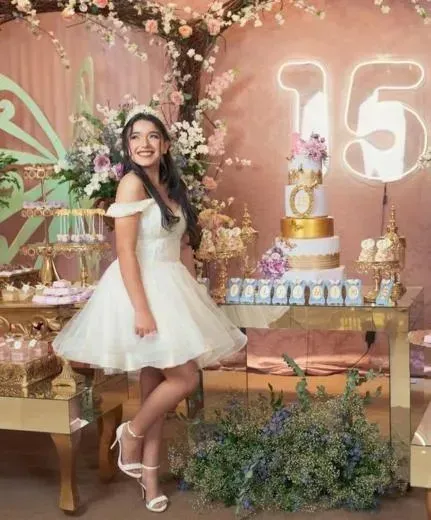 Woman in white dress posing at a pink floral 15th birthday party with cake and balloon decorations