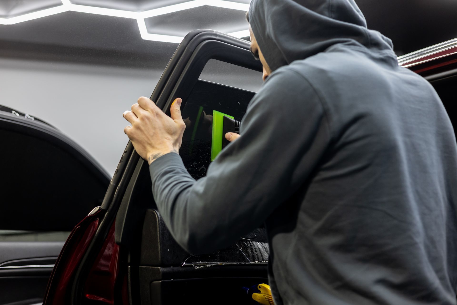 A man in a hoodie is applying window tinting to a car window.