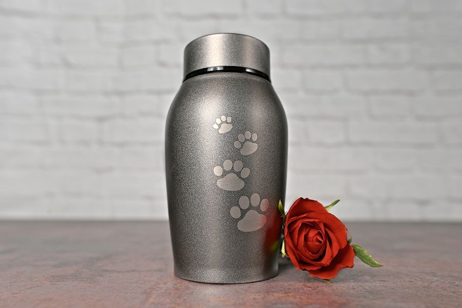 A metal urn with paw prints on it is sitting on a table next to a red rose.