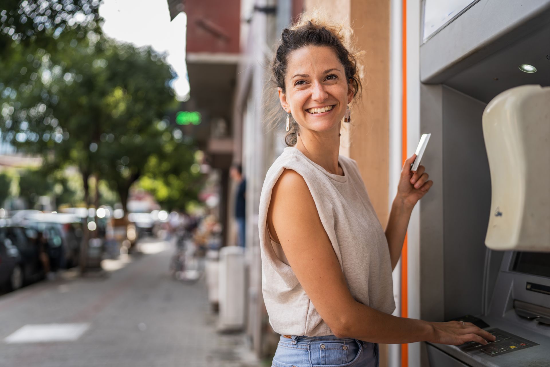 Woman smiling, using ATM on city street; holding card, typing with machine.