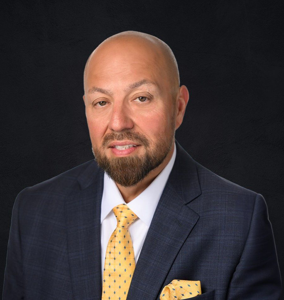 Bald man in a suit with a yellow tie and beard, smiling against a black background.