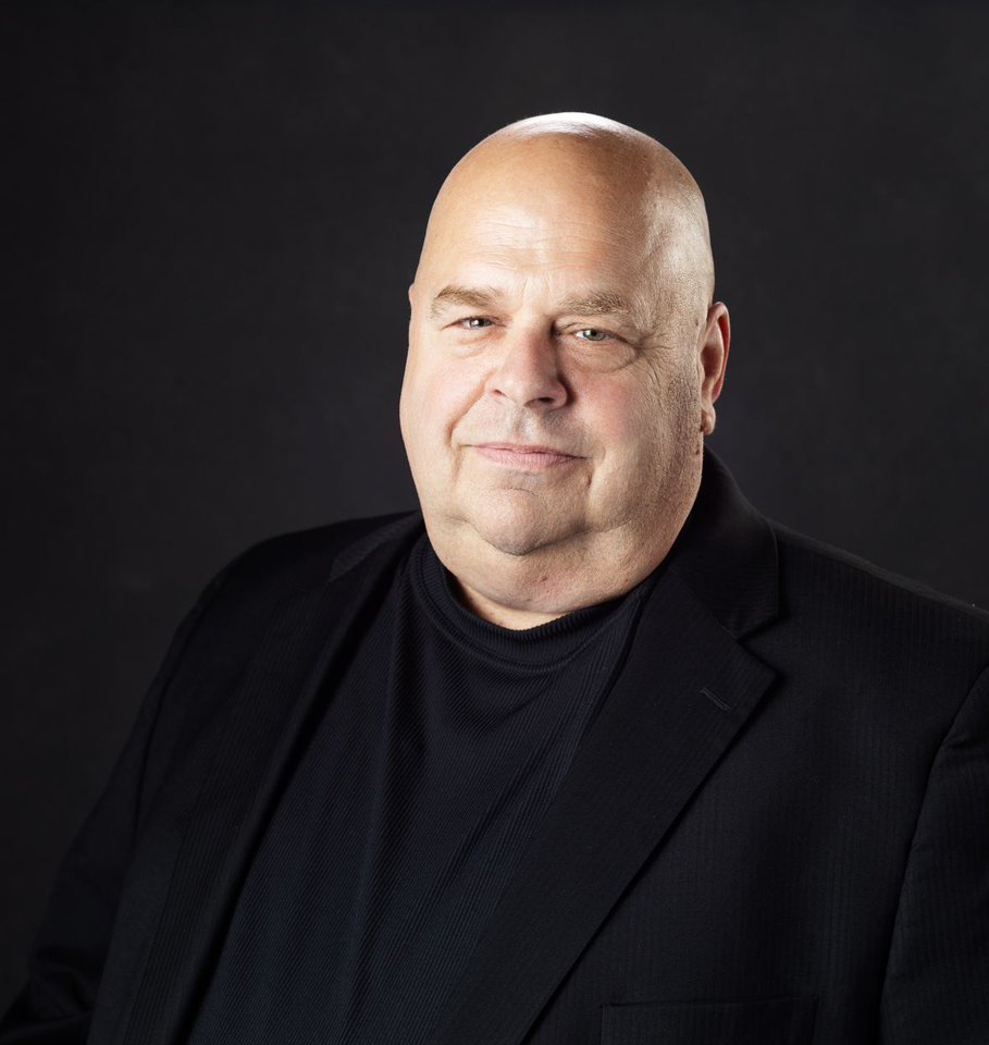 Bald man in a black jacket and sweater, against a dark background, looking forward.